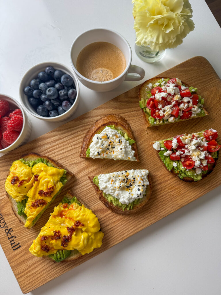 Three avocado toasts on a wooden board: soft egg with chili oil, cottage cheese with everything bagel, and tomato feta with hot honey