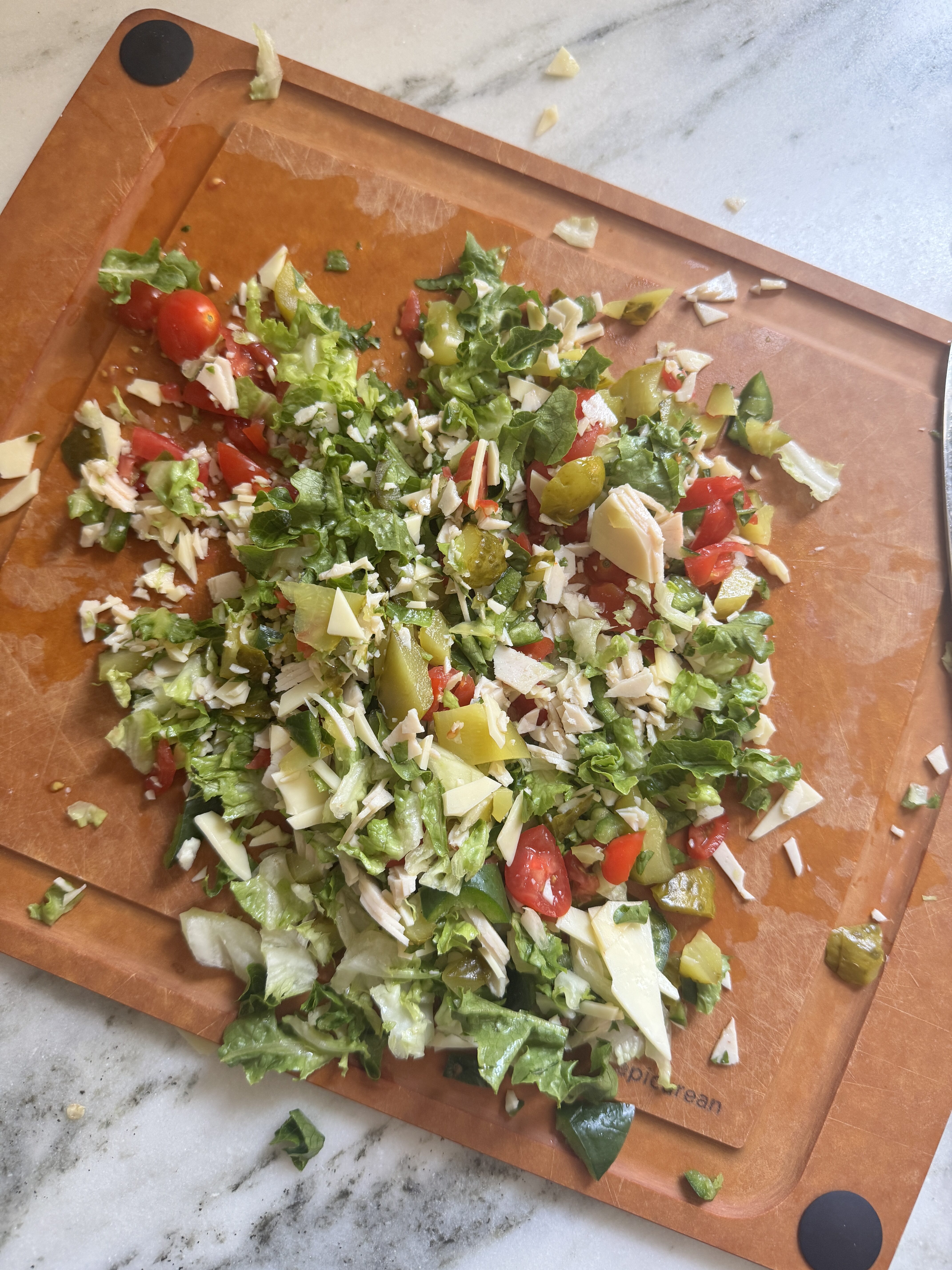 Freshly chopped sub salad ingredients spread across a wooden cutting board including lettuce, cherry tomatoes, turkey, cheddar cheese, pickle, and jalapeño