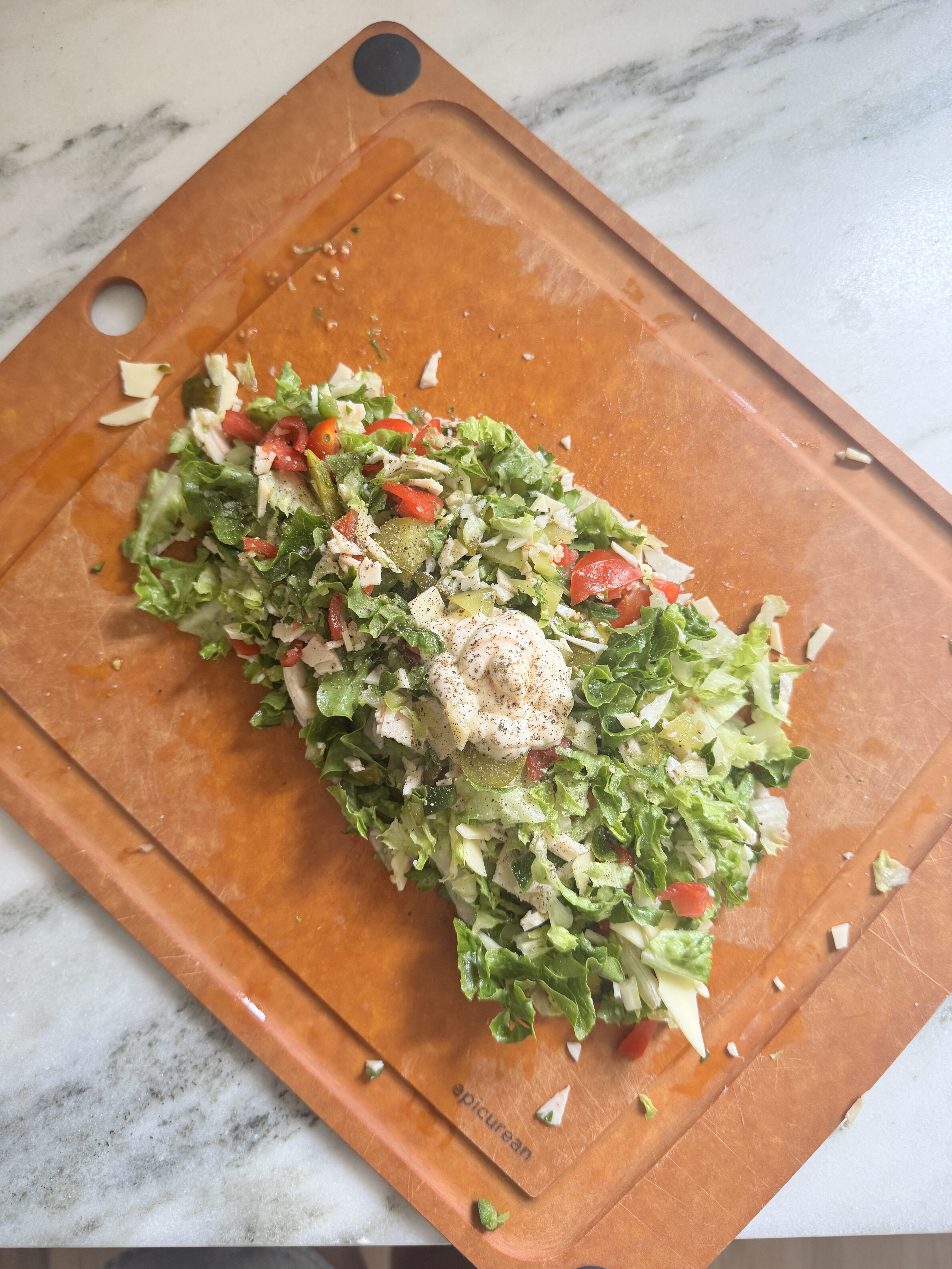 Chopped sub salad ingredients on a wooden cutting board with a dollop of mayo dressing on top, ready to be tossed, overhead view