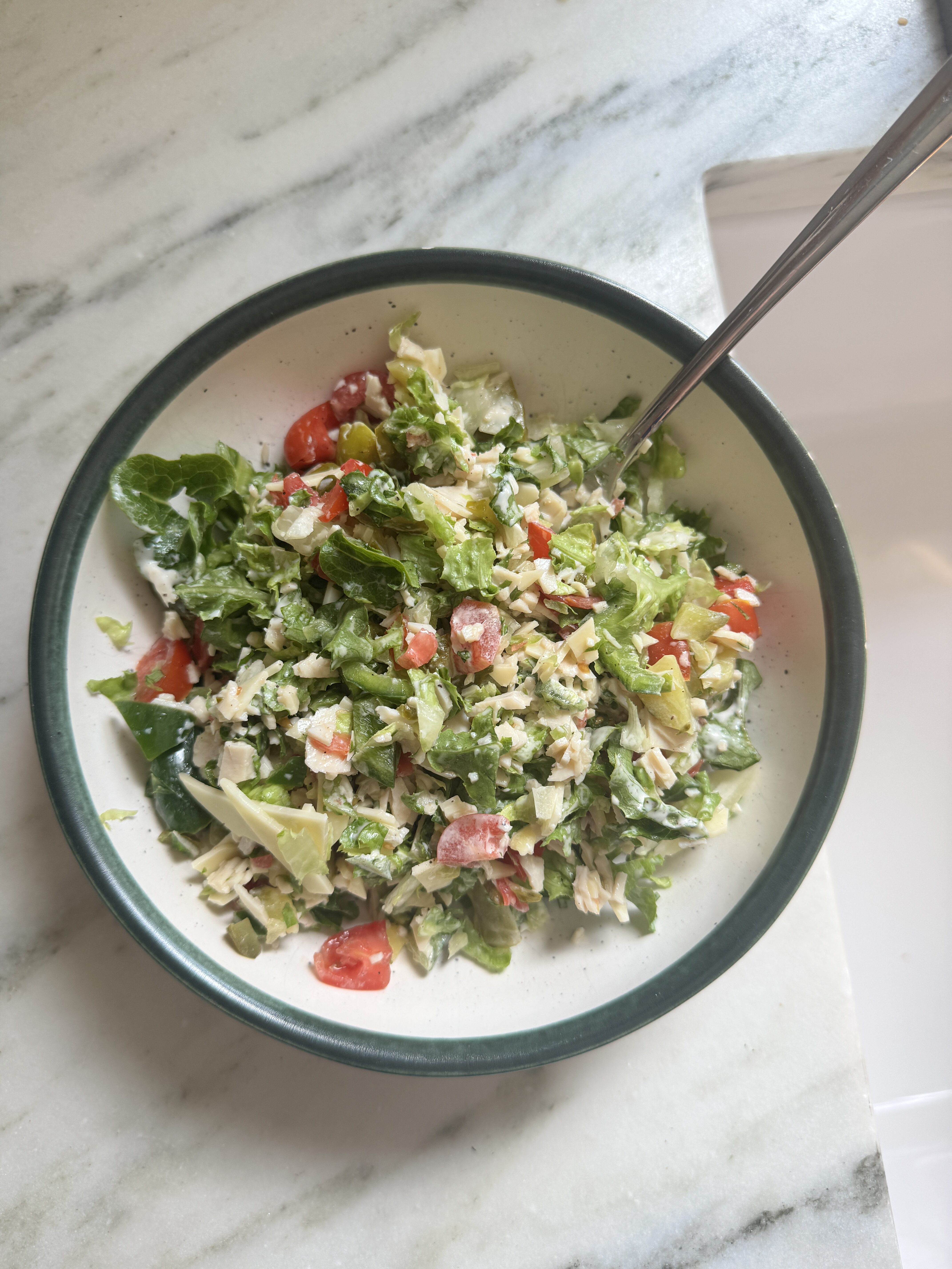 Chopped sub salad in a white bowl with green rim, showing finely chopped lettuce, cherry tomatoes, turkey, cheddar cheese, and jalapeño tossed in creamy mayo dressing, overhead view on marble surface