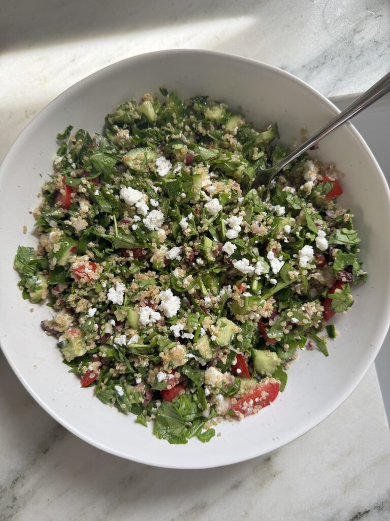 Mediterranean quinoa salad in a white serving bowl topped with feta, kalamata olives, cherry tomatoes, cucumber, and fresh parsley.