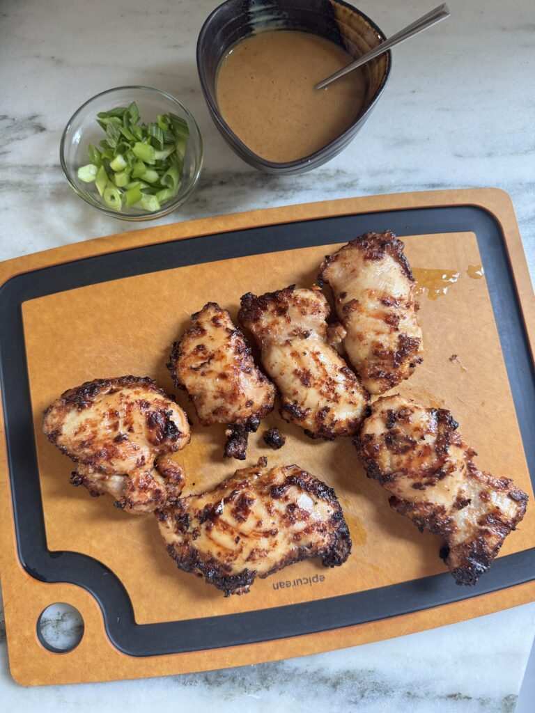 Peanut chicken thighs on a cutting board with a bowl of peanut sauce and green onion