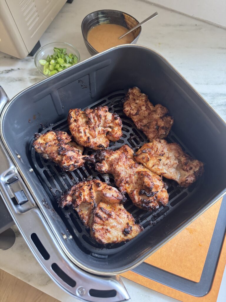 Cooked peanut chicken thighs in an air fryer basket with peanut sauce and green onion