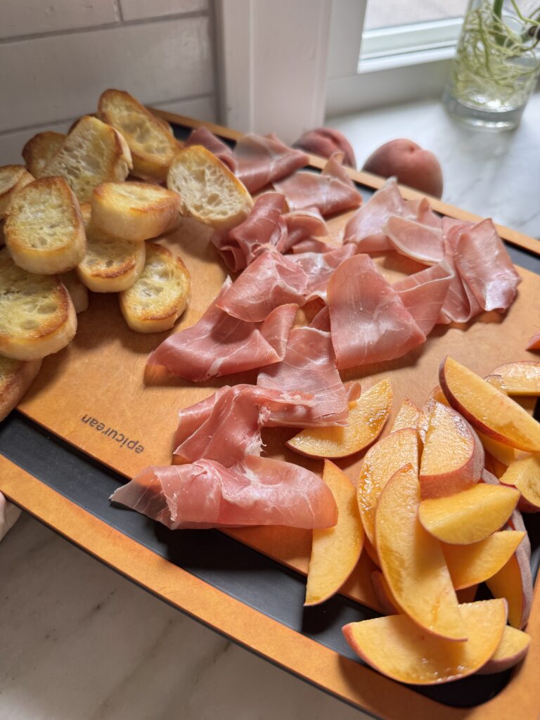 Toasted crostini, prosciutto, and sliced peaches laid out on a board to assemble