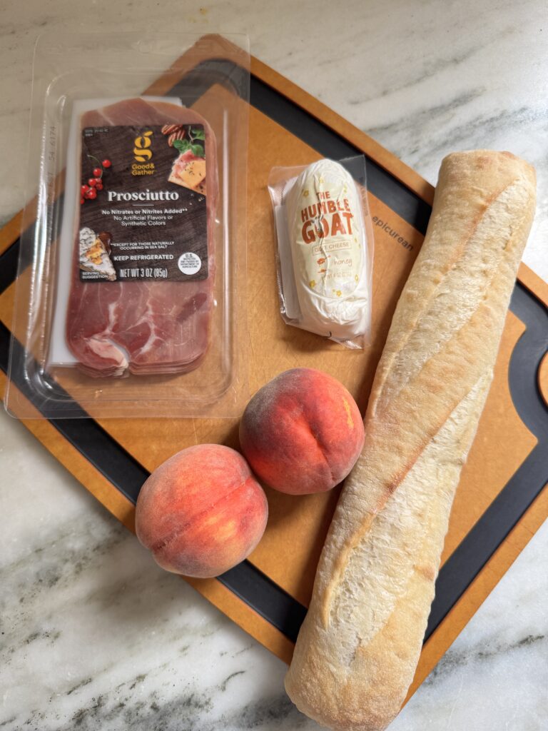 Prosciutto, goat cheese, fresh peaches, and a baguette on a cutting board