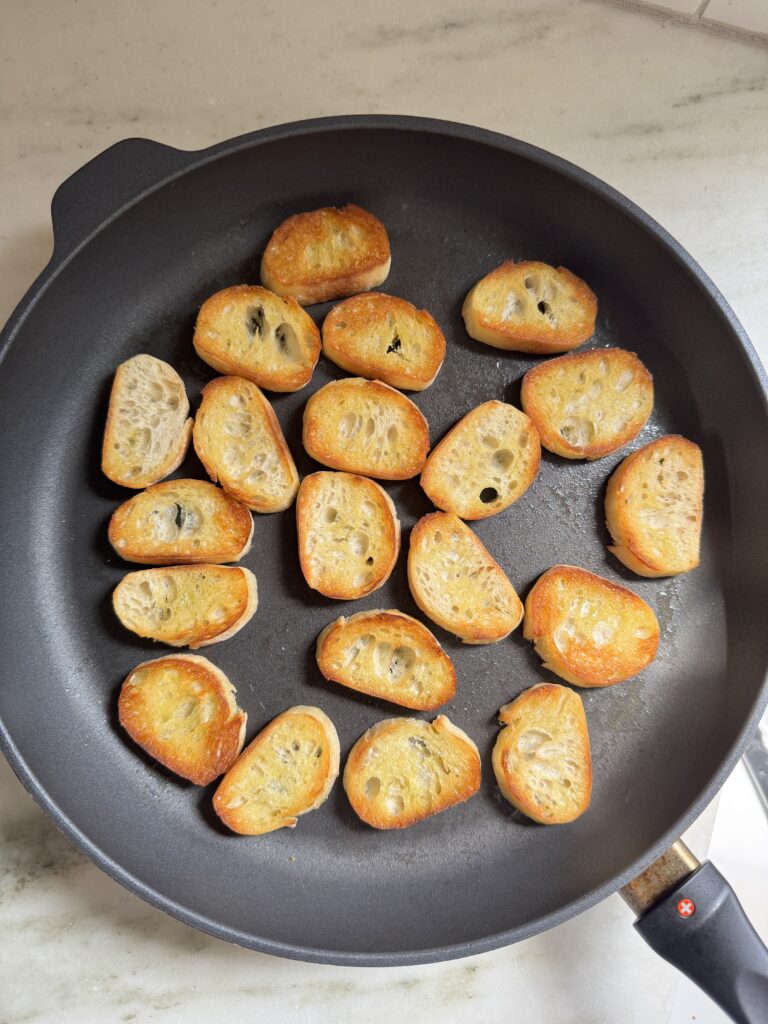 Baguette slices toasting in a skillet for peach prosciutto crostini