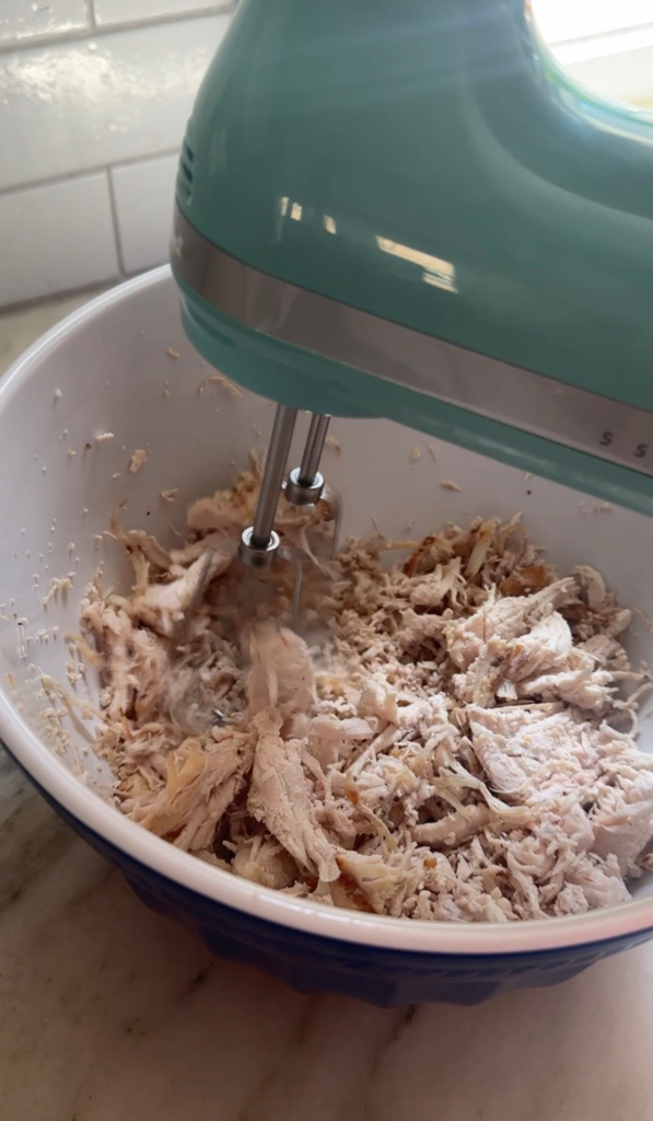 Shredding cooked chicken with a KitchenAid hand mixer in a bowl for pepperoncini chicken salad