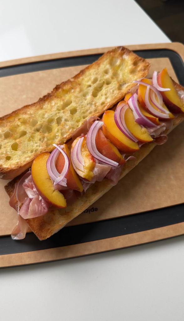 Sliced peaches and red onion added over prosciutto on a toasted baguette