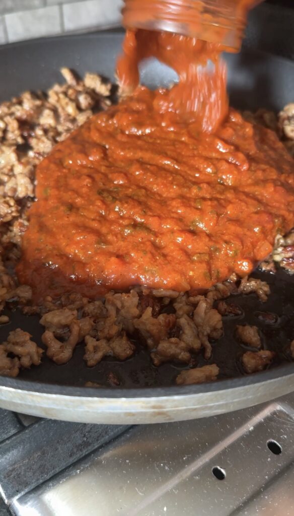Pouring marinara sauce over browned Italian sausage in a skillet