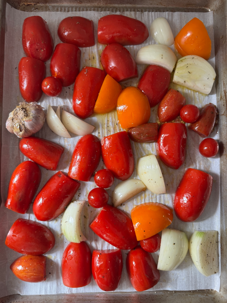 Raw Roma tomatoes, onion, and a garlic bulb on a baking sheet before roasting