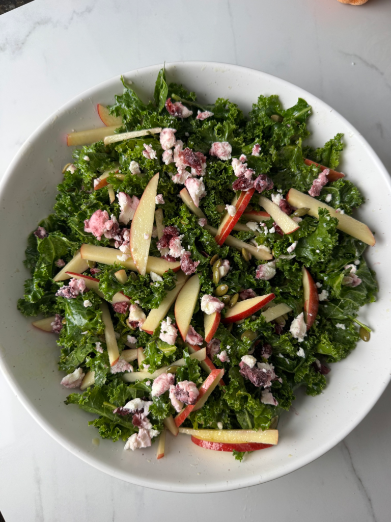 A bowl of kale salad with apple, cranberry goat cheese, and pepitas